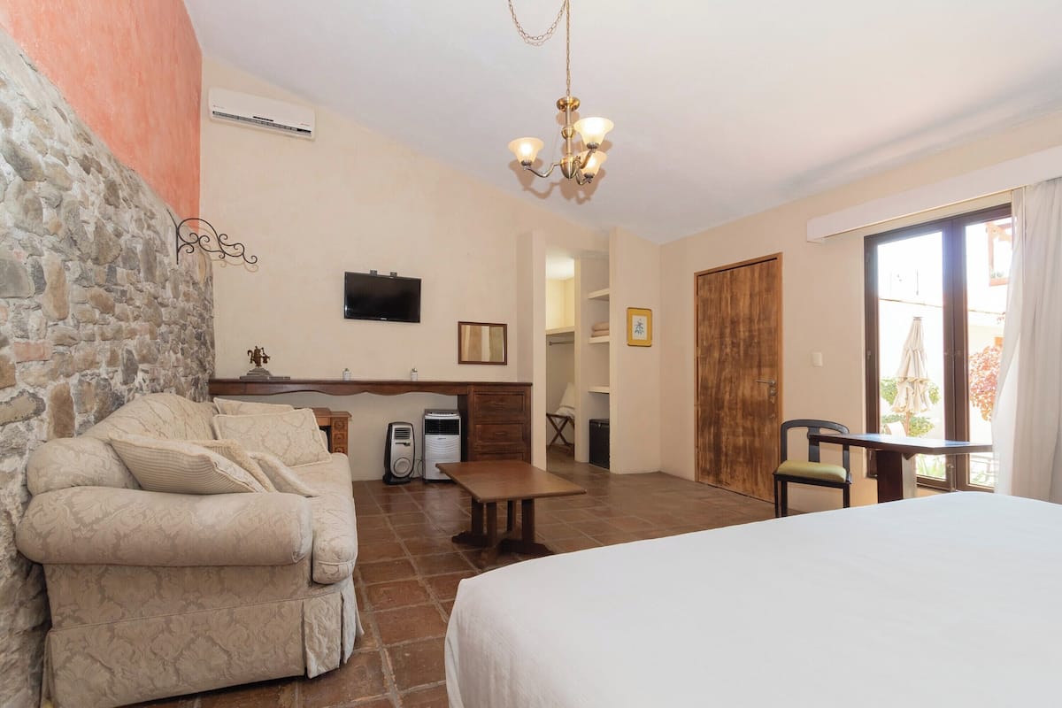 A room with a bed, a sofa, a desk, a chair, a TV, and a door leading to another room.
