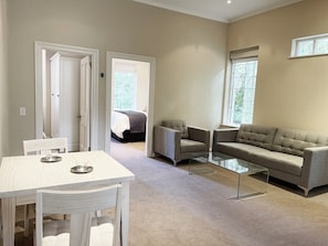 Un salon moderne avec une table à manger blanche, deux fauteuils, un canapé et une table basse en verre. Deux portes donnent sur une chambre, et une fenêtre apporte de la lumière naturelle.