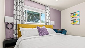 A bedroom with a bed, a nightstand, a window, and a wall with abstract art. - Big bang in a little boxx (Warren)