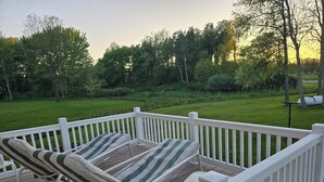 Una terraza con dos sillones de descanso y una mesita, rodeada por una baranda blanca, con vistas a un césped cuidado y árboles.