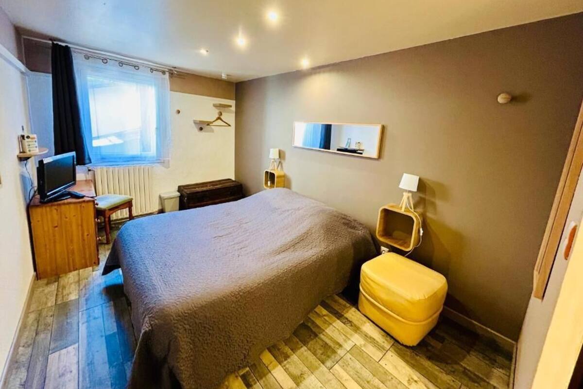 Une chambre avec un grand lit, une table de chevet, une chaise, une petite table avec une lampe, un miroir et une fenêtre avec des rideaux.