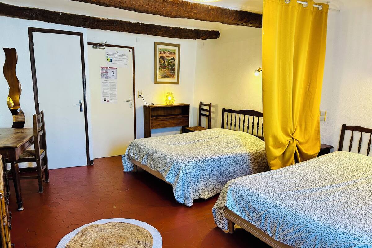 Une chambre avec deux lits, une chaise en bois, une petite table, une lampe et une porte sur laquelle est fixé un tableau d’affichage.