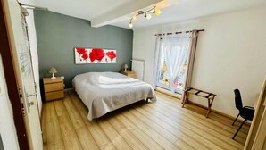 Un dormitorio con cama, mesita de noche, lámpara, silla, ventana con cortinas y un cuadro en la pared.
