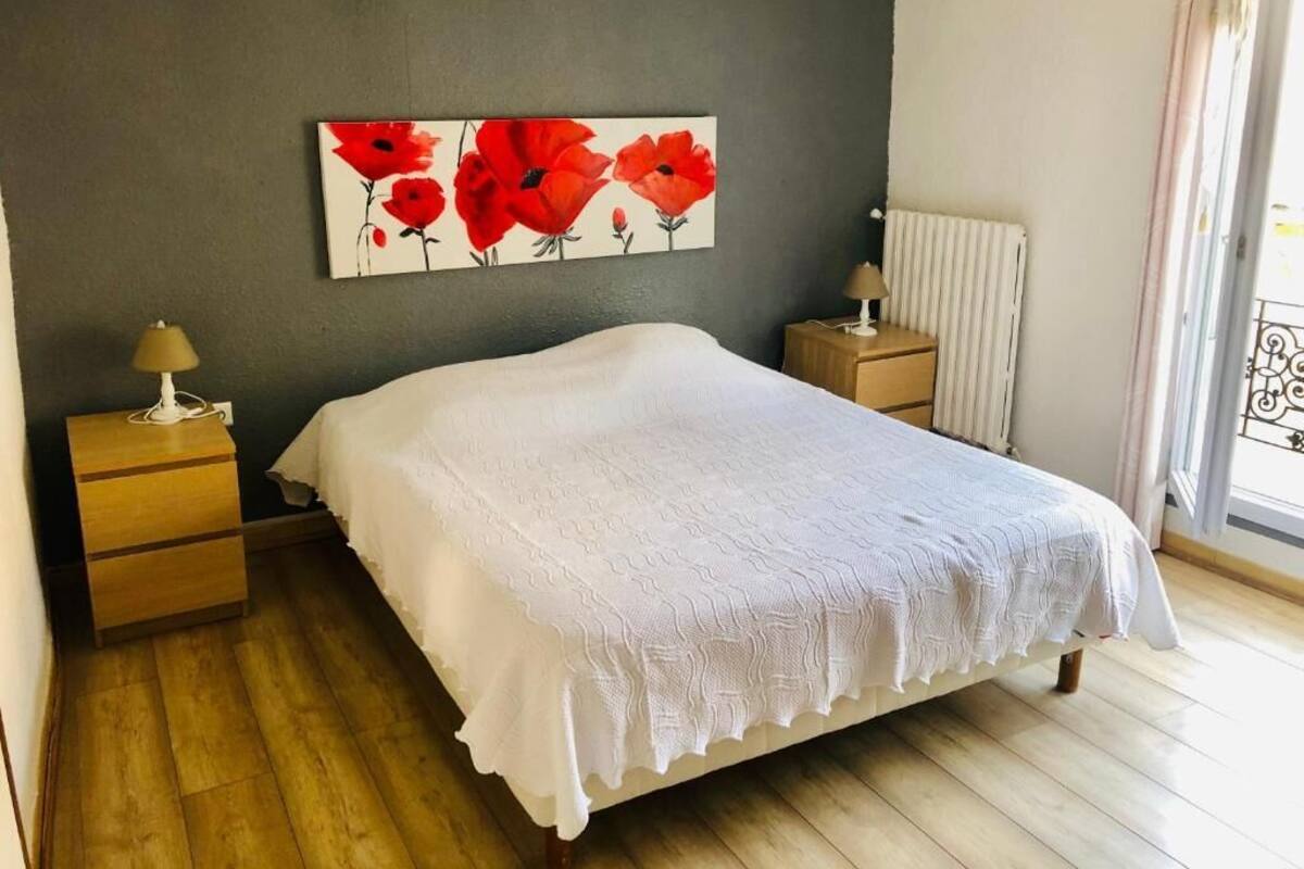 Une chambre à coucher avec un lit, une table de chevet, une lampe, un radiateur et une fenêtre avec des rideaux.