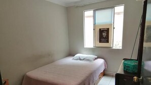 Quarto pequeno com cama de solteiro, janela, ventilador de teto e porta.