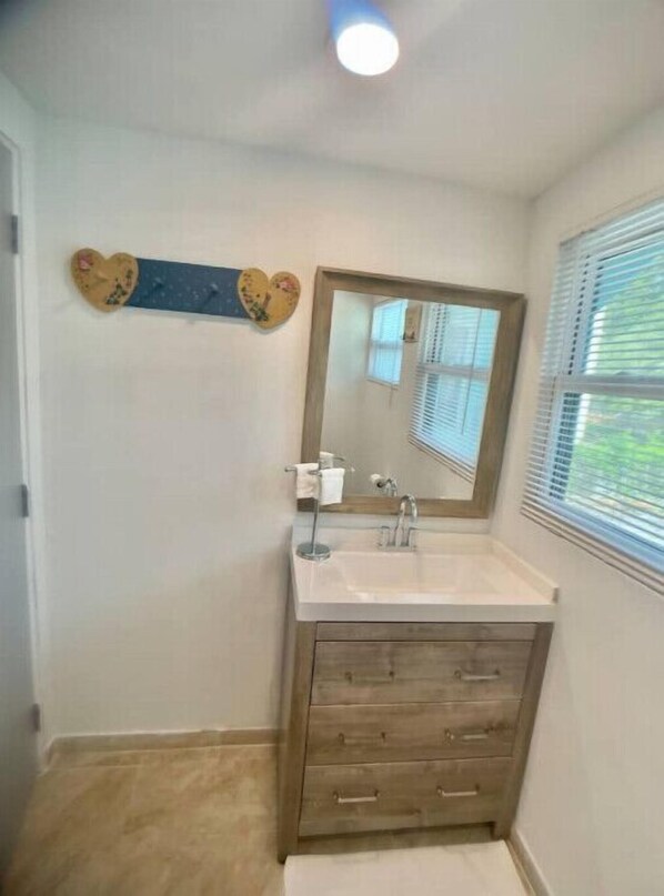 A bathroom with a wooden vanity, a mirror, and a window with blinds.
