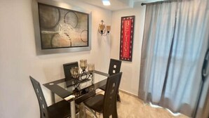 A dining area with a glass table, black chairs, a framed abstract painting, a wall-mounted light fixture, and a 'BELIEVE' sign.