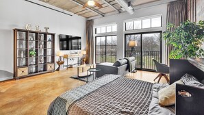 Ein geräumiges Wohnzimmer mit einem großen Bett, einem Sofa, einem Schreibtisch und einem Bücherregal. Große Fenster und ein Deckenventilator sind vorhanden.