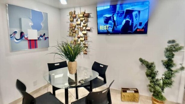 Una sala de reuniones moderna con una mesa de vidrio, sillas negras, una planta en maceta, un televisor de pared mostrando una película y arte abstracto en las paredes.