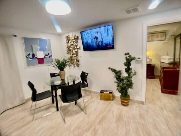 Un comedor moderno con una mesa de vidrio y sillas negras, un televisor montado en la pared y una planta en maceta.