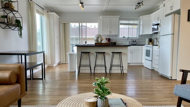 A modern kitchen with white cabinets, a central island with stools, and a dining area with a leather couch and a round wooden coffee table. - Spacious 4-BR Home near Jasper! (Hinton)