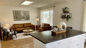 A modern living room with a brown sofa, a coffee table, and a dining area with a white cabinet and a dark countertop. - Spacious 4-BR Home near Jasper! (Hinton)