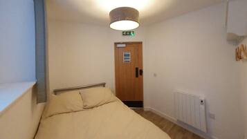 A narrow hallway with a bed on the left, a wooden door at the end, and a radiator on the right.