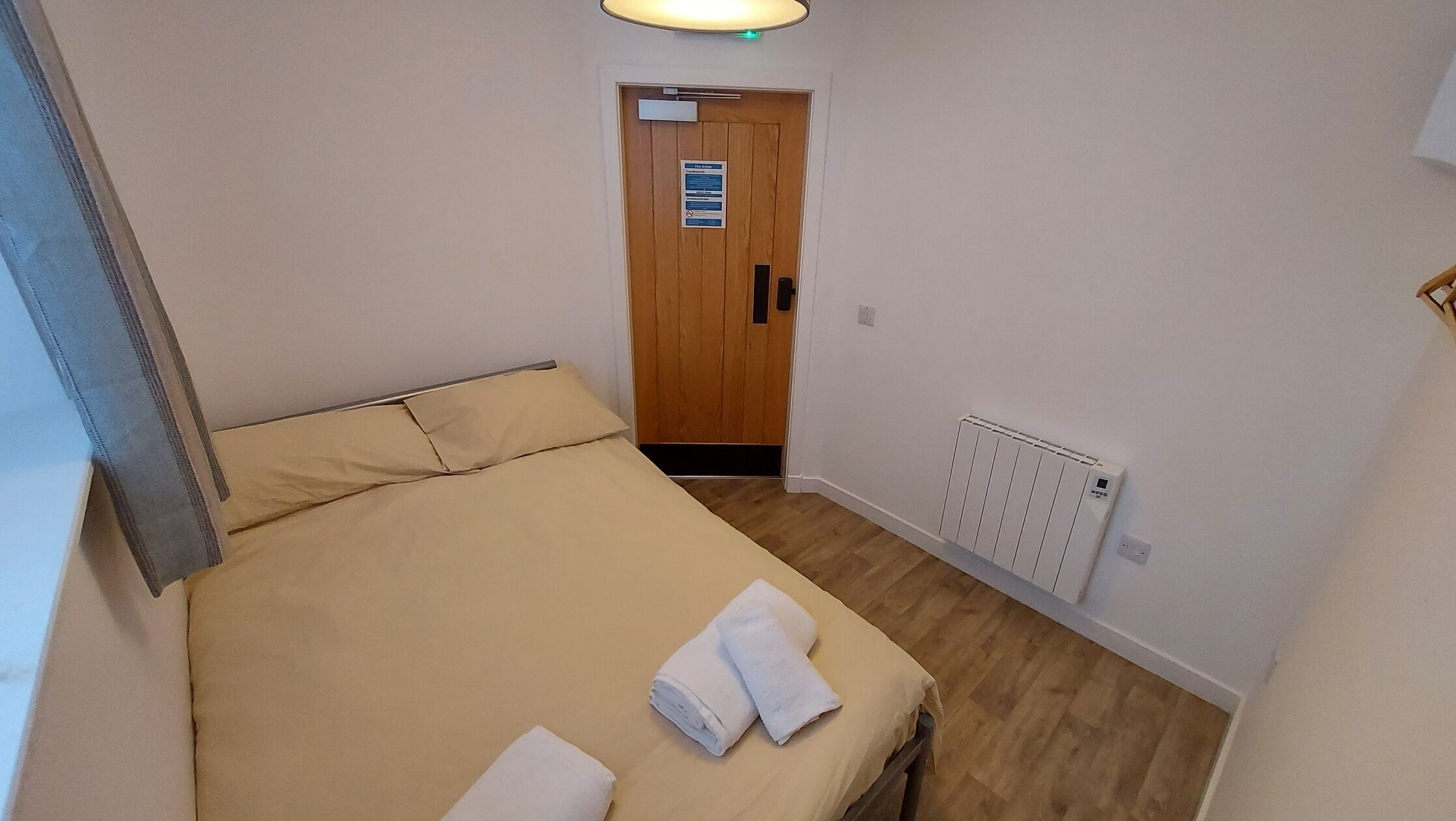 A small, narrow hotel room with a single bed, a wooden door, a radiator, and a light fixture above the door.