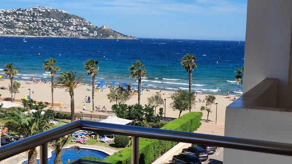 A beach view from a balcony with palm trees, a pool, and a view of the sea and a hill with buildings.