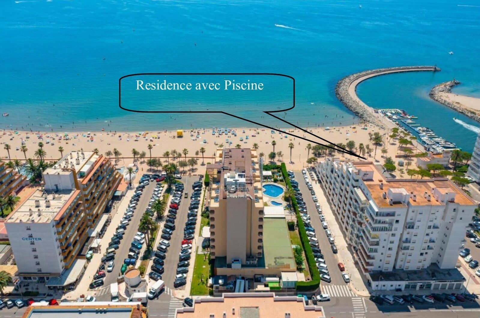 A hotel with a swimming pool, located near a beach with a curved pier and numerous boats.