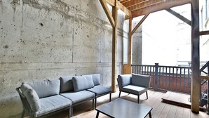 Un interno moderno con una parete in cemento, un soffitto in legno, un divano grigio, un tavolino e un balcone con ringhiera in metallo.