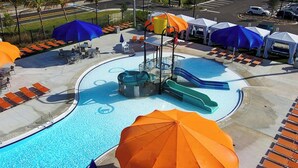 Een zwembad met ligbedden en parasols, omgeven door een geplaveid gebied met een waterglijbaan en een kleine brug.
