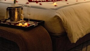 A hotel room with a bed, a bedside table with a bucket and glasses, a nightstand with a lamp, and a framed abstract painting on the wall.