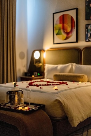 A hotel room with a bed, a bedside table with a bucket and glasses, a nightstand with a lamp, and a framed abstract painting on the wall.