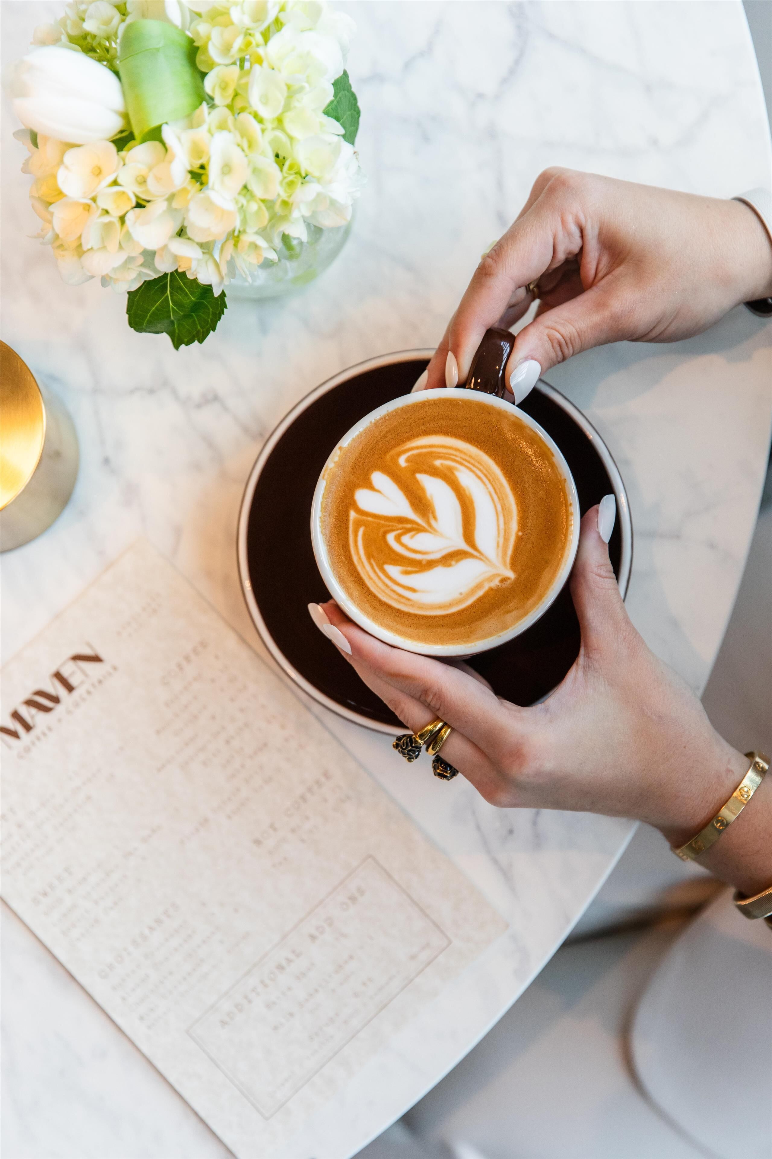 Een persoon houdt een koffiekop vast met latte-art, een menukaart en een bloevas op een marmeren tafel.
