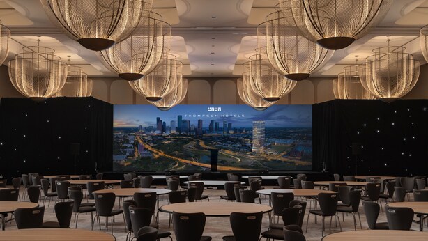 A conference room with a large screen displaying a cityscape, multiple tables and chairs, and modern chandeliers.