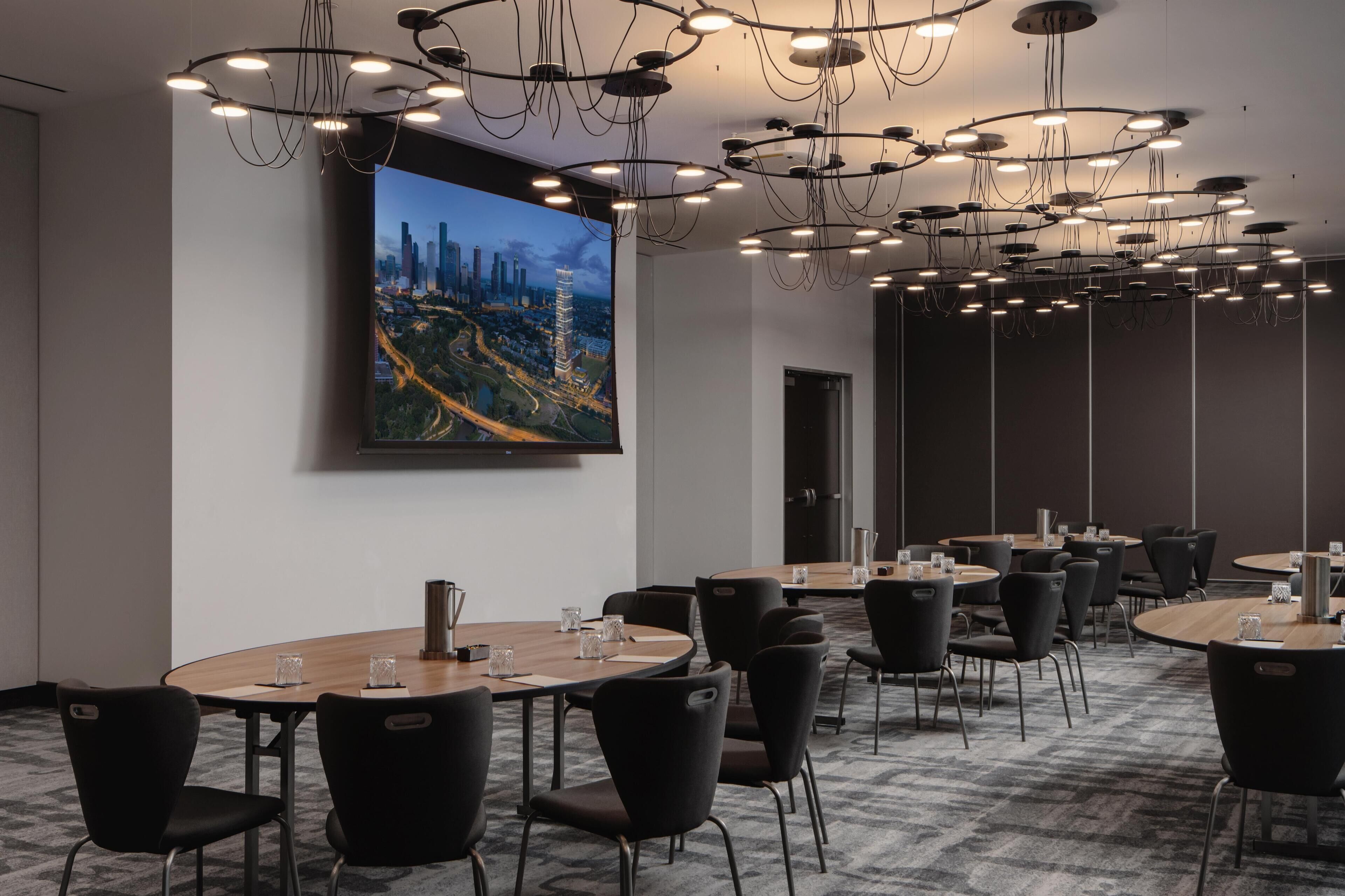 Une salle de conférence dotée de plusieurs tables et chaises, d’un grand tableau représentant un paysage urbain et d’un lustre moderne.