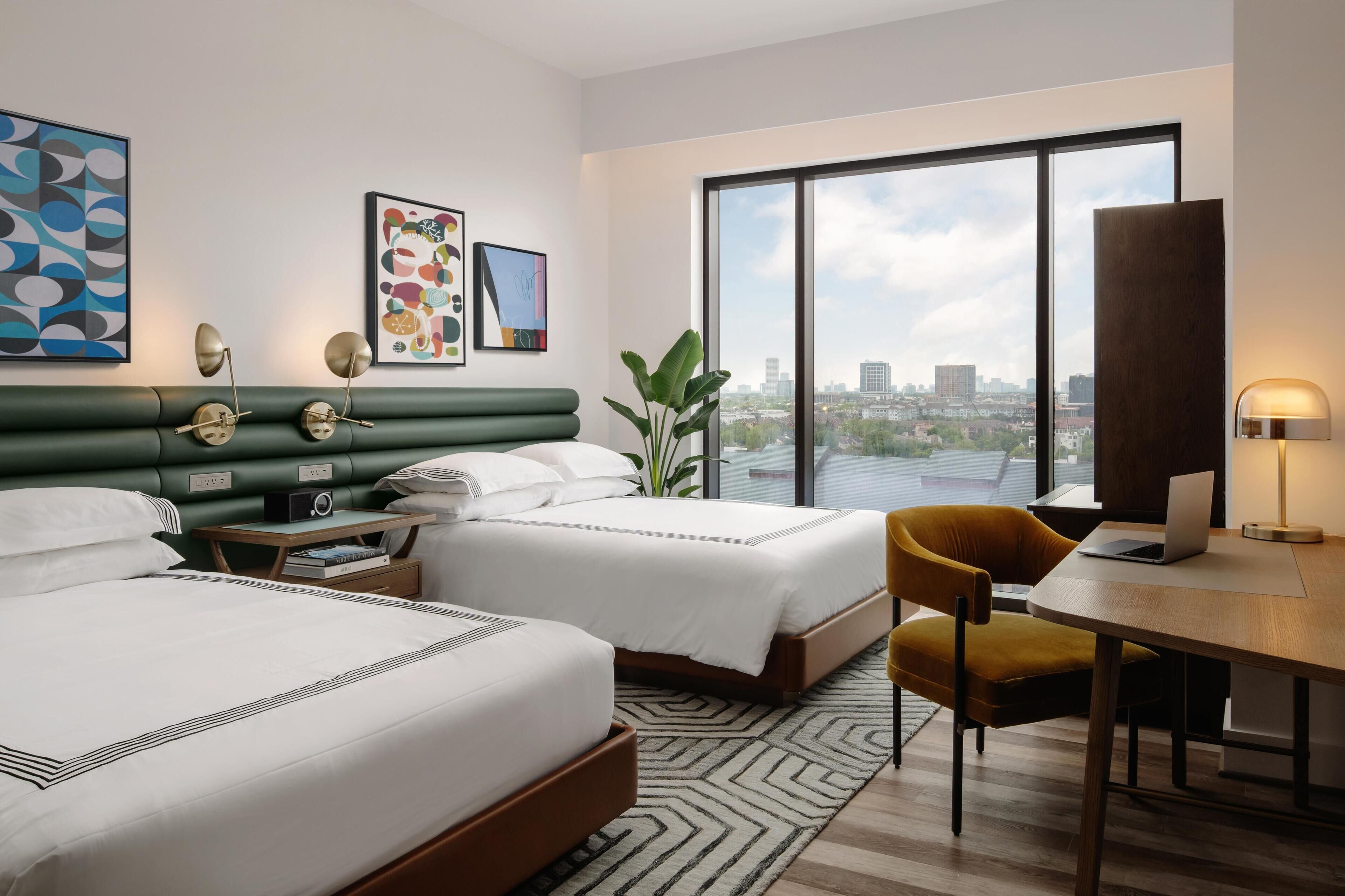 A hotel room with two beds, a desk, a chair, and a large window offering a city view.