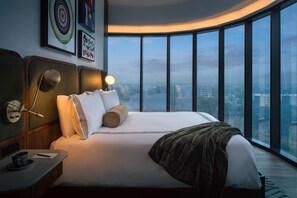 A modern hotel room with a large bed, a bedside table with a lamp, a view of the city through large windows, and artwork on the walls. - Thompson Houston, by Hyatt (Houston)