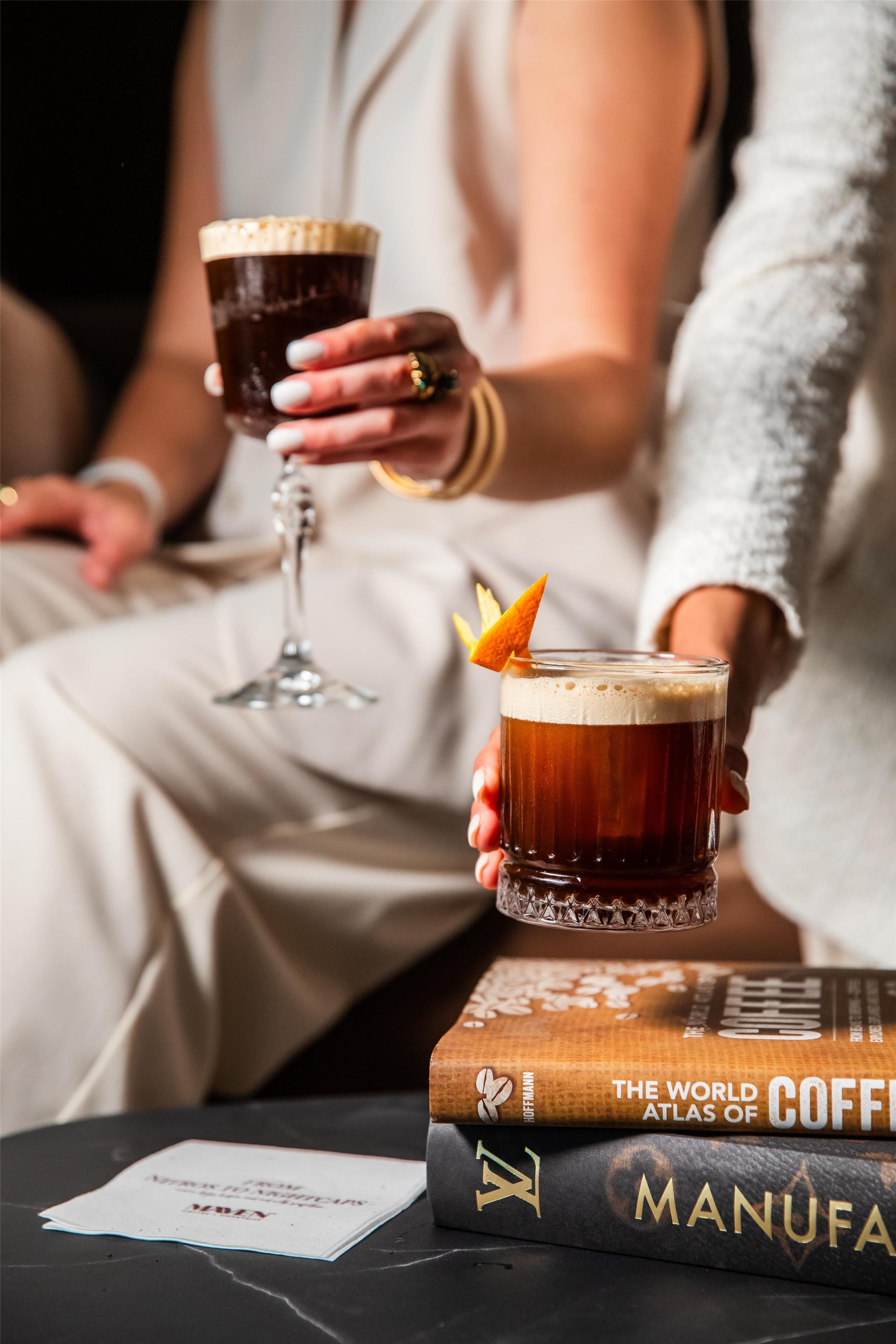 A person holding a glass of beer with a lemon slice garnish, seated next to a stack of books titled 'The World Atlas of Coffee' and 'Manu.'
