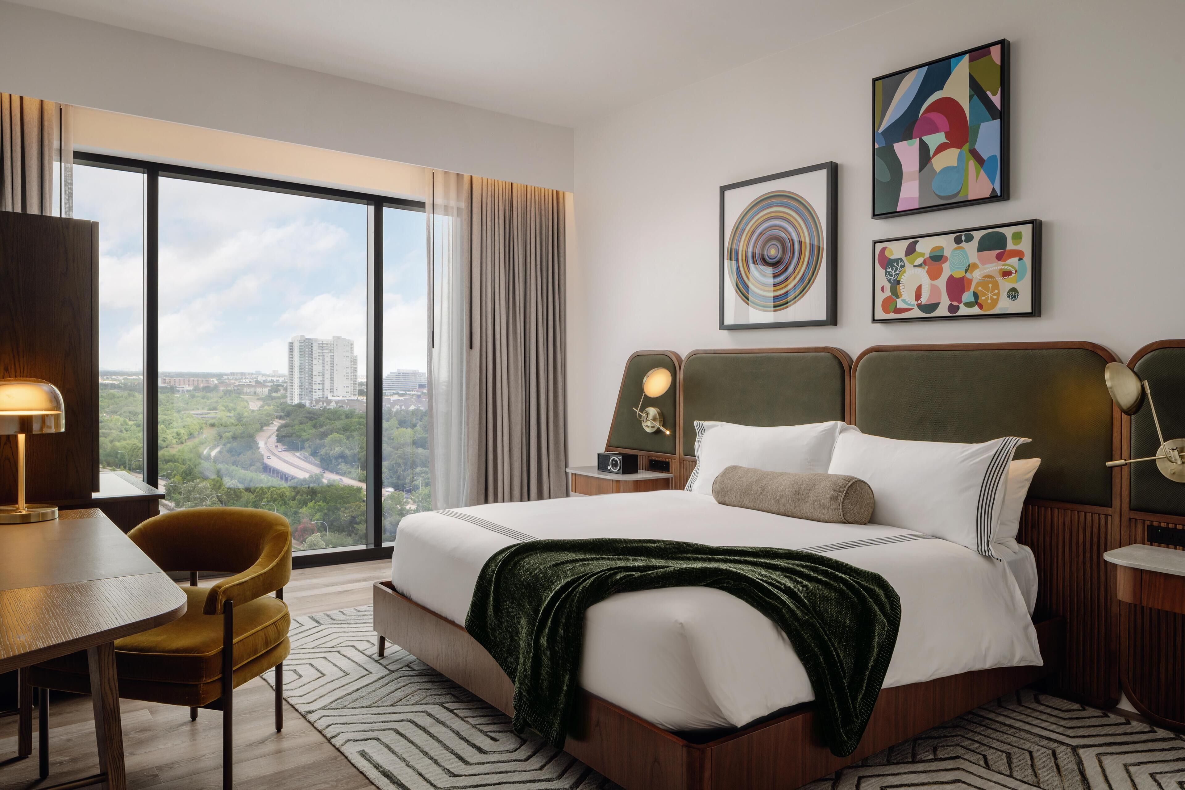 A hotel room with a large window, a bed with a green throw, a desk with a lamp, and two wall-mounted artworks.