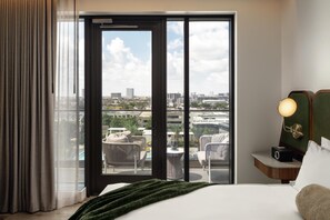 A hotel room with a bed, a nightstand with a lamp, a view of the city through a large window, and a balcony with outdoor furniture. - Thompson Houston, by Hyatt (Houston)