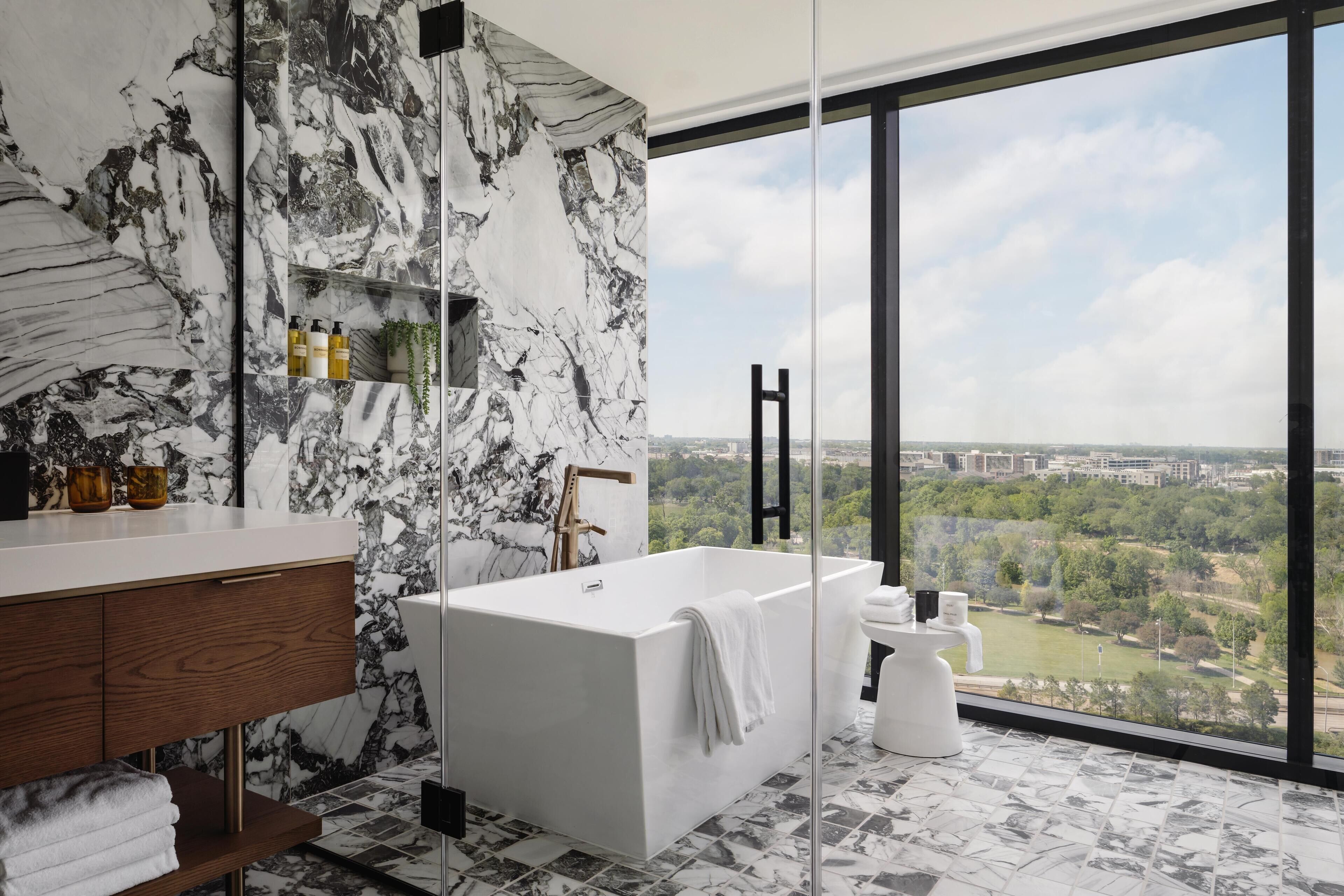Un baño moderno con pared de mármol, una bañera, un amplio ventanal con vista a la ciudad y un mueble de tocador de madera.