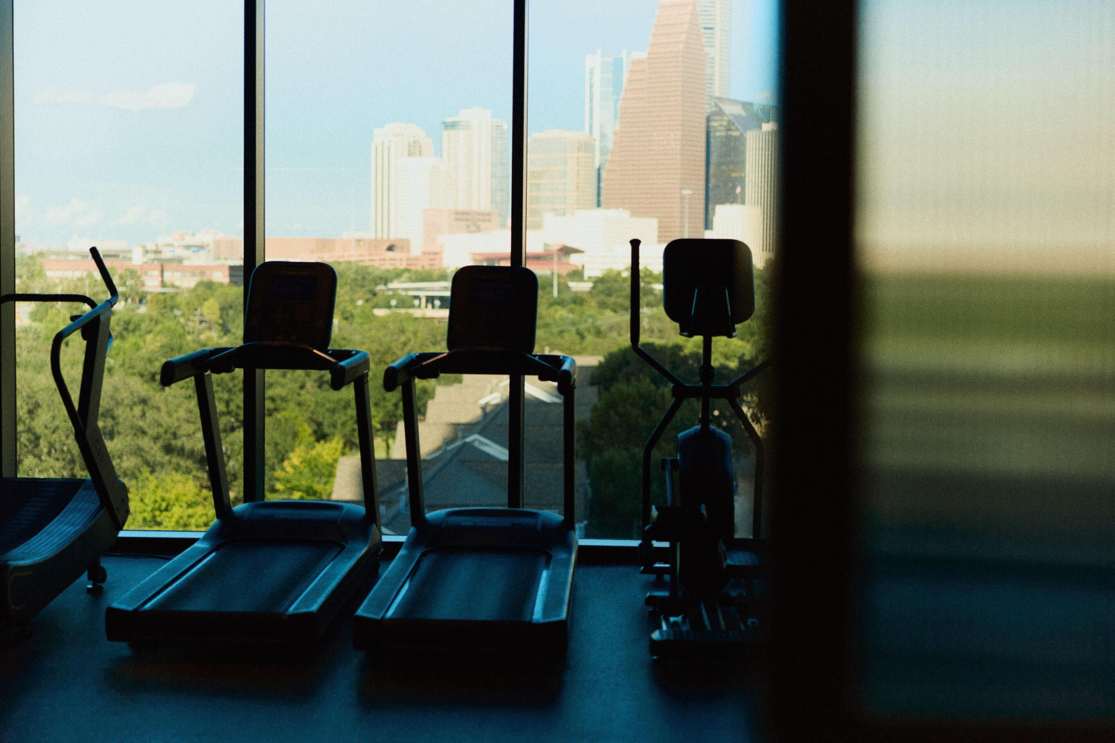 An indoor gym with exercise equipment and a view of a city skyline through large windows.