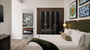 A modern hotel room with a bed, a desk, a TV, and artwork on the walls.