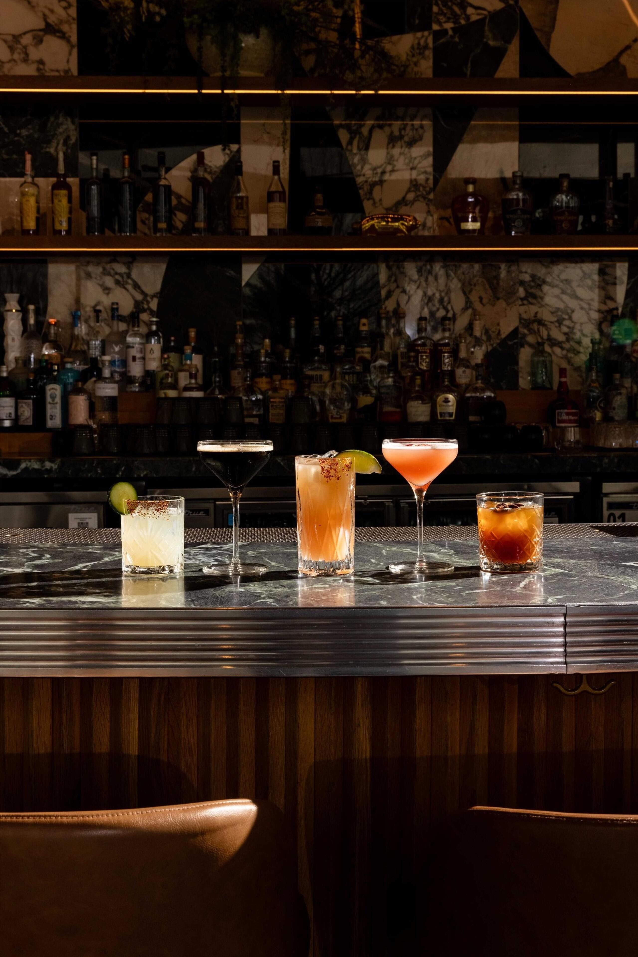 A bar with four different cocktails on the counter, a variety of bottles in the background, and a marble wall.