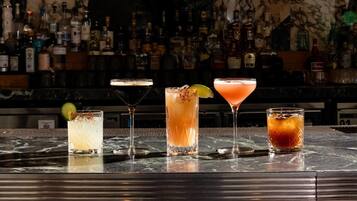 A bar with four different cocktails on the counter, a variety of bottles in the background, and a marble wall.
