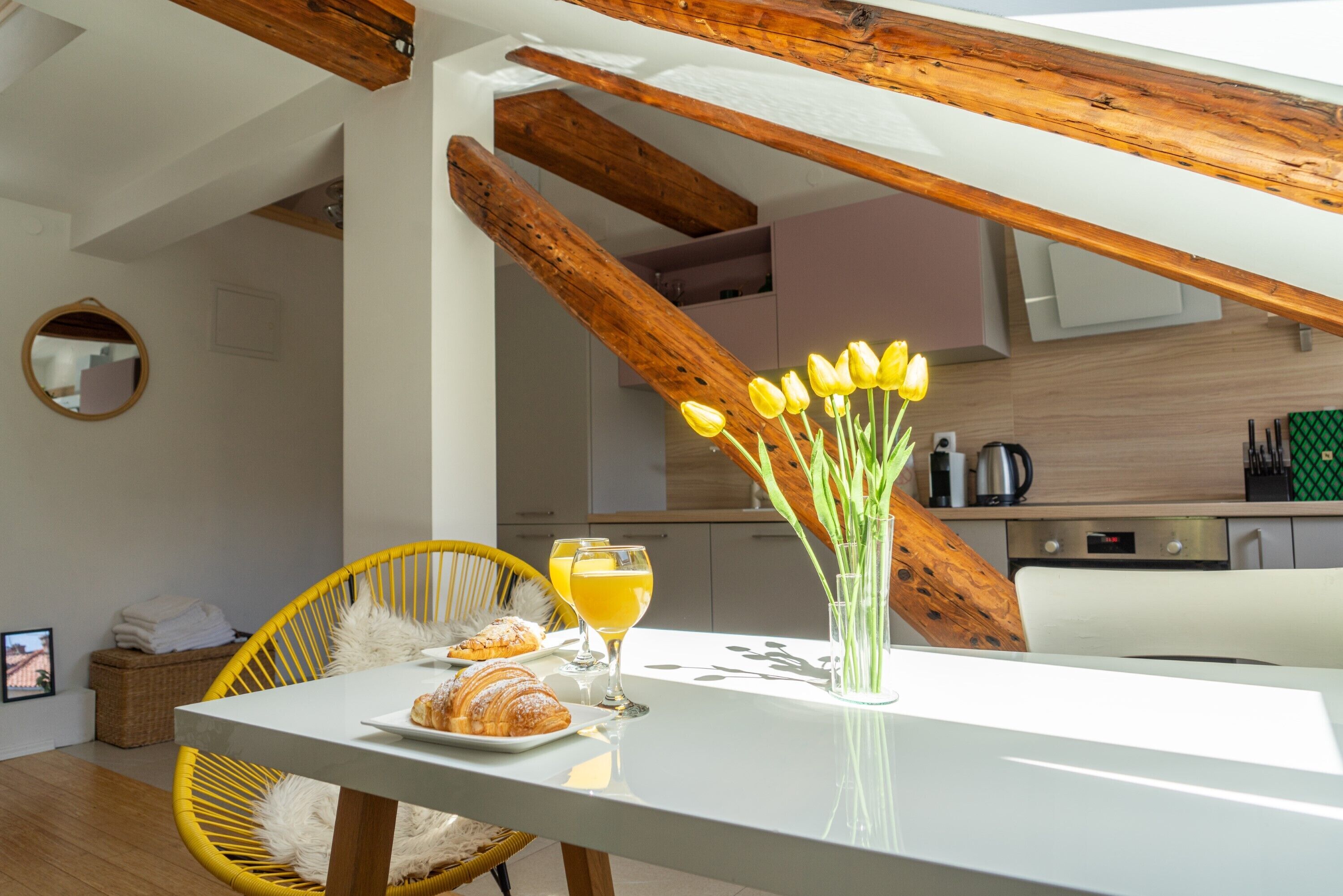 Una cocina moderna con vigas de madera, una mesa de comedor con un florero y una preparación para el desayuno con cruasanes y jugo de naranja.