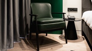 Een moderne hotelkamer met een groen bekleden fauteuil, een klein bijzettafeltje met een telefoon, en een bed met een witte hoofdbord.