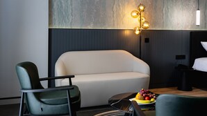 Een moderne hotelkamer met een gebogen bed, twee groene fauteuils, een ronde salontafel met fruit, en een bijzondere wandlamp.