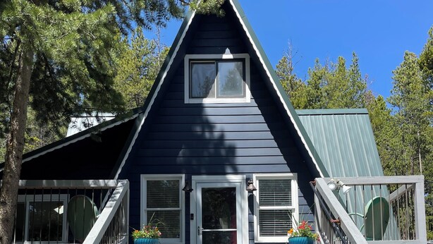 A two-story house with a steeply pitched roof, a front porch with a railing, and a set of stairs leading to the entrance. - Mountain Bliss is a quiet mountain retreat, sleeps 6, hot tub, pet friendly (Evergreen)