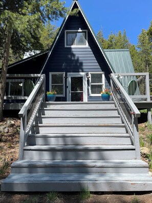 A two-story house with a steeply pitched roof, a front porch with a railing, and a set of stairs leading to the entrance. - Mountain Bliss is a quiet mountain retreat, sleeps 6, hot tub, pet friendly (Evergreen)