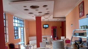 Interior de restaurante con suelo a cuadros, mesas y sillas blancas, televisión en la pared y un pilar rojo.
