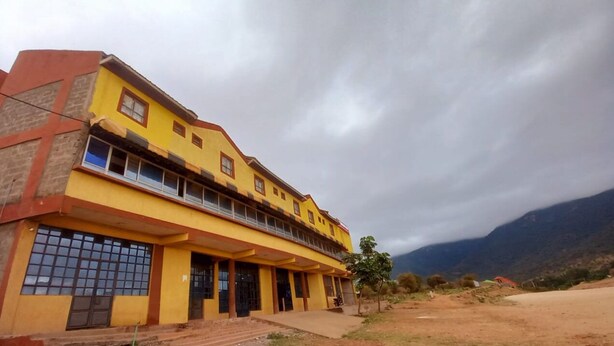 Un edificio amarillo con múltiples ventanas y un balcón, rodeado de un paisaje montañoso.