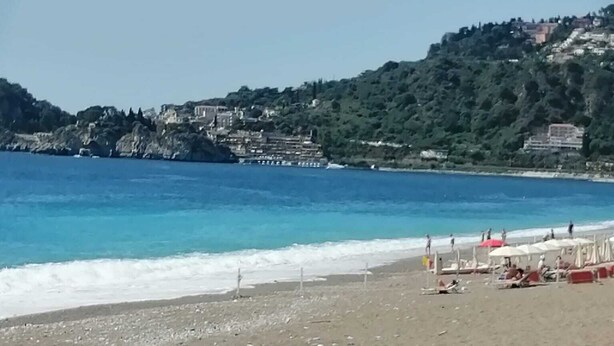 Ein Strand mit klarem, blauem Wasser, einem sandigen Ufer und Gebäuden, die sich an einen Hügel schmiegen.