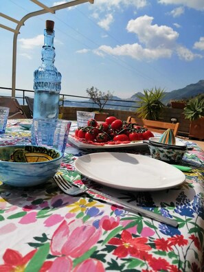 A table set outdoors with a view of mountains, a bottle of water, a plate of tomatoes, and a bowl. - Sicily and Sun (Letojanni)