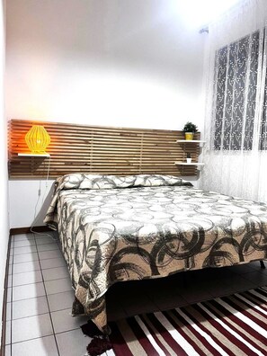A bedroom with a bed, a headboard, a bedside table, a lamp, a potted plant, and a window with curtains. - Sicily and Sun (Letojanni)