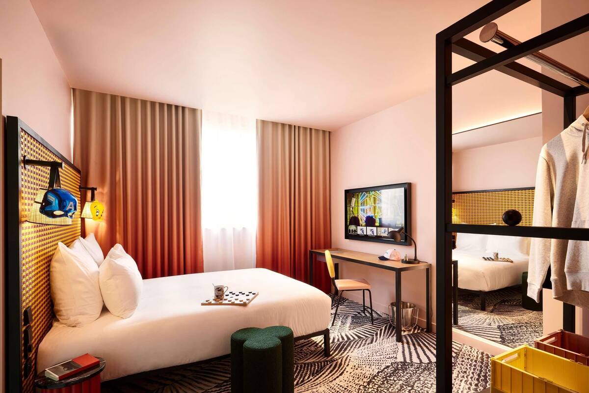 A hotel room with a bed, desk, TV, and a mirror reflecting the room.