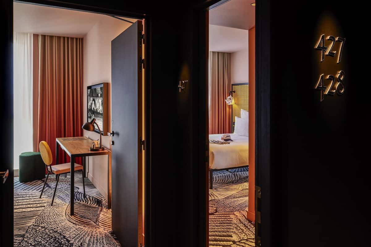 A hotel room with a bed, a desk, a chair, a painting, and a patterned carpet.