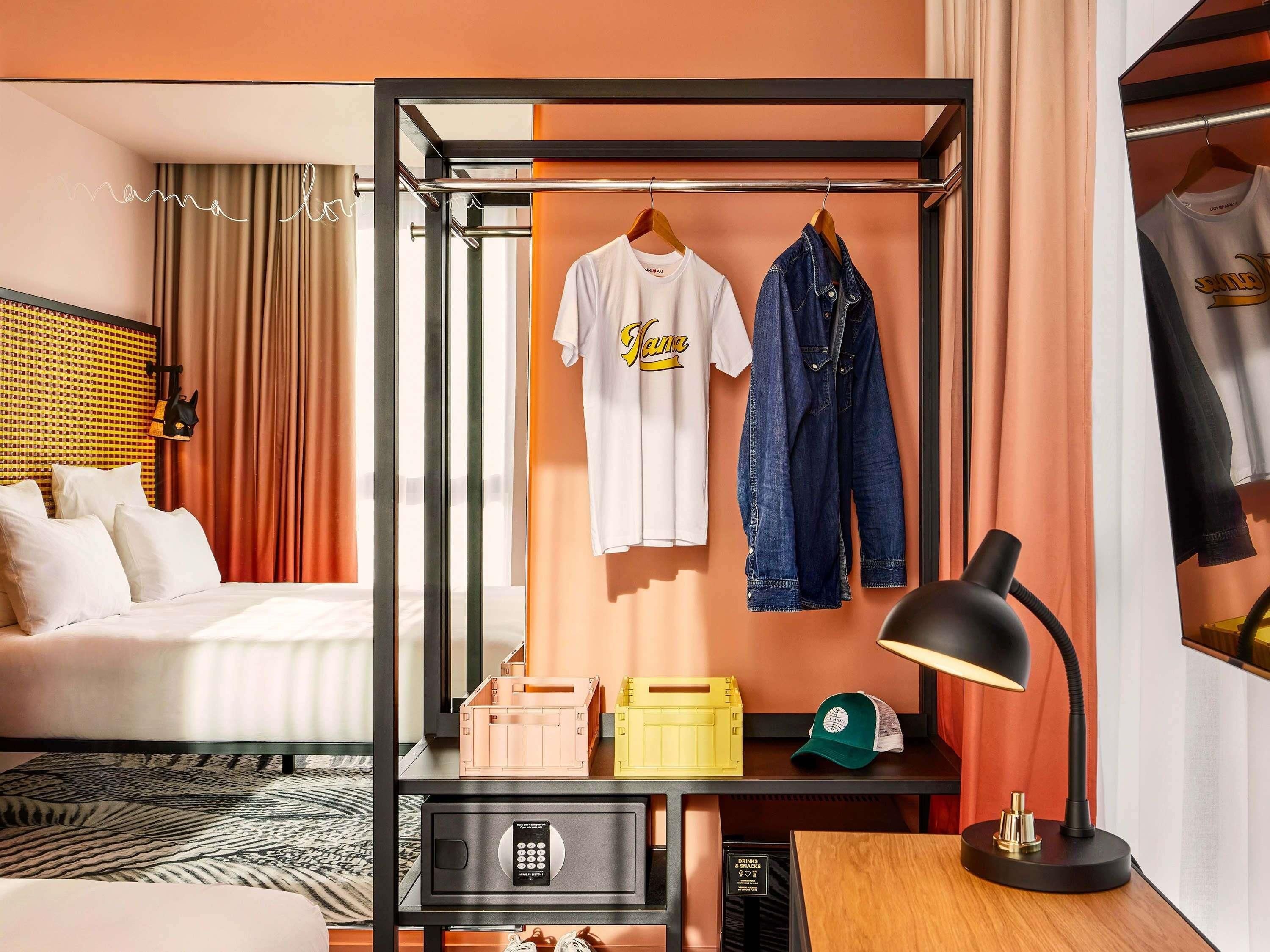 Een hotelkamer met een bed, een bureau met een lamp, een kledingrek met een t-shirt en jeans, en een klein kluisje.